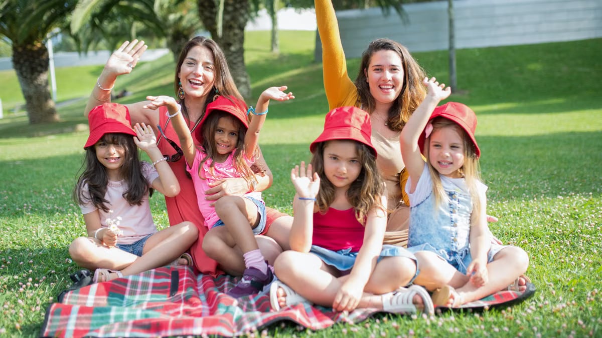 Two women and four girls enjoy a sunny picnic in Portugal, waving happily in the park.